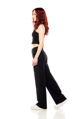 Charming young redhead woman posing in studio in black short blouse and black trousers on a white background. Side view
