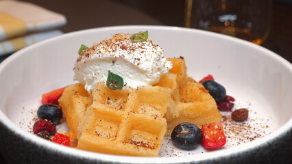 Delicious golden waffles topped with whipped cream, fresh blueberries, strawberries, and chocolate shavings in white bowl, perfect for breakfast or brunch ideas. Healthy Eating and Lifestyle.