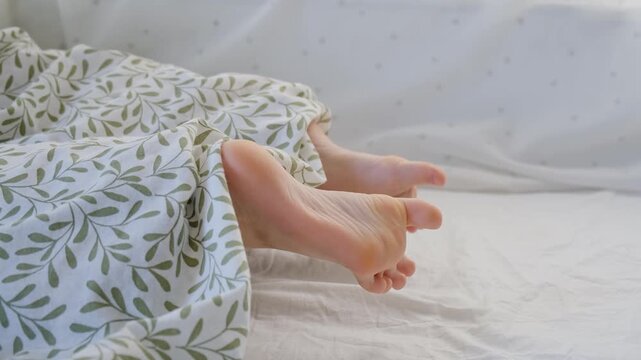 The bare feet of child sticks out from under the blanket and stretching after morning wake up. Sleeping on stomach. Lying in soft fresh bed linen and mattress. Daytime sleep. Happy childhood concept