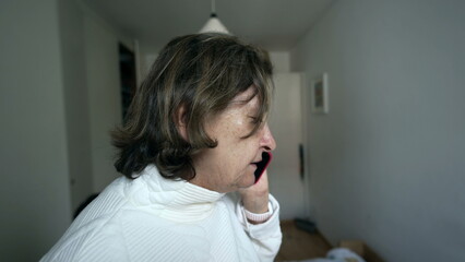 Older woman talking on phone, wearing glasses, white sweater, indoors, serious expression, home setting, natural light, modern apartment, communication, focused, close-up view, everyday life