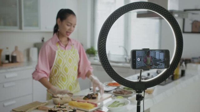 Young Asian girl blogging about kimbap cooking on smartphone and ring light while preparing dinner at home kitchen