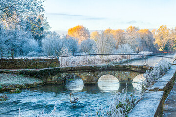 The old stone bridge over the River Coln and hoar frost on Rack Isle at sunset in the Cotswold village of Bibury, Gloucestershire, England UK