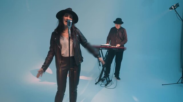 A vocalist in a black leather jacket and hat,smiling and singing with eyes open,stands at a vintage microphone with hands moving up and down A keyboardist works a red sampler against a blue background