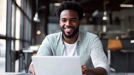 Fototapeta premium Confident and Connected: A young Black professional radiates success, smiling brightly while working on his laptop in a modern office. 