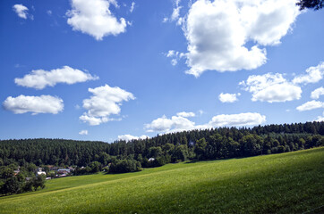 Landscape on a slope. A grassy landscape with a small village in a valley. Blue sky with white clouds. Forests in the background. Nature with a village on a sunny summer day.