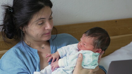 Mother gazing lovingly at newborn baby while holding and working on laptop, highlighting the balance between work and parenthood, dedication in a cozy home setting