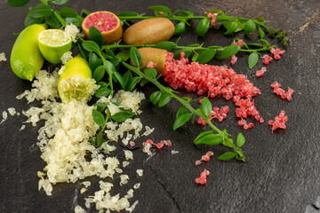 Ripe yellow and burgundy oblong fruits and delicious pink and white citrus caviar on the black slate slab decorated with a green plants sprig. Outlandish Australian finger lime plant indoor growing