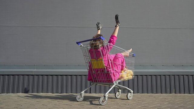 Beautiful woman in pink costume having fun in the grocery cart under the supermarket outdoors. Women racing shopping trolleys on the street background. Positive female going crazy