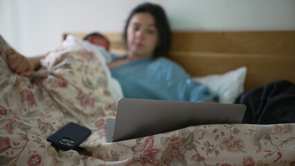 Mother in bed with newborn baby sleeping on her chest, using a laptop, combining work and care in a...