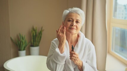 Woman applying natural drops cosmetics oil to face skin. Mature adult old woman applying nourishing anti age serum with pipette doing daily beauty routine. Senior woman gets kin scare facial treatment