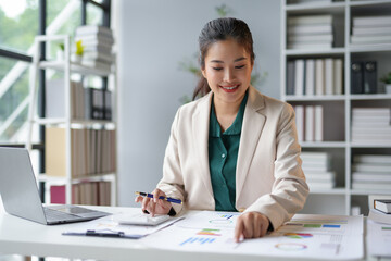 Smiling asian businesswoman works confidently in a modern office, analyzing financial data on her laptop surrounded by charts and graphs