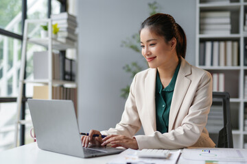 Young asian businesswoman is smiling and working on a laptop while sitting at her desk in a modern...