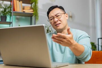 Displeased sad Asian man use laptop notebook typing browsing working, loses becoming surprised sudden lottery results bad news fortune loss game fail computer virus. Mature guy at home room on couch