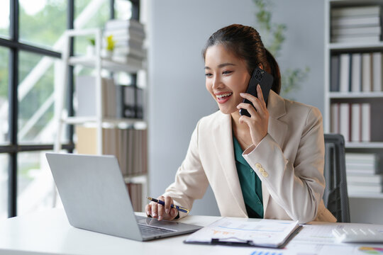 Smiling asian businesswoman talking on phone and using laptop while working at office desk