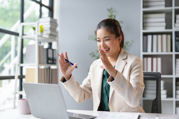 Businesswoman is having a video call on her laptop and waving at the camera while sitting at her...