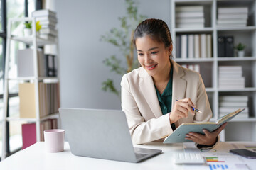 Smiling asian businesswoman holding pen and taking notes in notebook while having video call on laptop at office