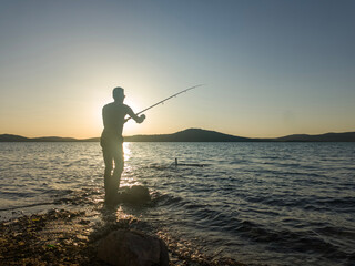 Fishing for pike, perch, carp. Fisherman with rod, spinning reel on sea or ocean. Man catching fish, pulling rod while fishing on sea, pond. Wild nature. The concept of rural getaway. Back view.