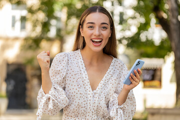 Oh my God Wow. Excited happy joyful winner young woman using smartphone looking at screen browsing celebrating win receive good message news. Caucasian lady with mobile phone in urban street outdoors