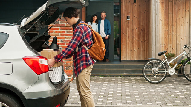 Happy son holding box with his things and positively looking straight at camera. In background mother and dad see their child joining road trip. Young teenager getting ready to drive to college.