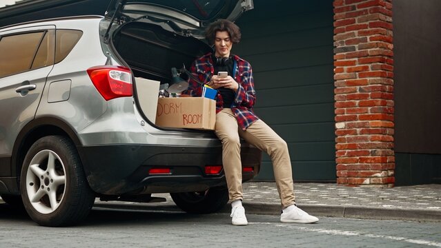 Young man entering college and loading cardboard boxes into car. Adult boy sitting on car trunk and using phone for texting messages to his friends. Teenager moving out of parent's home.