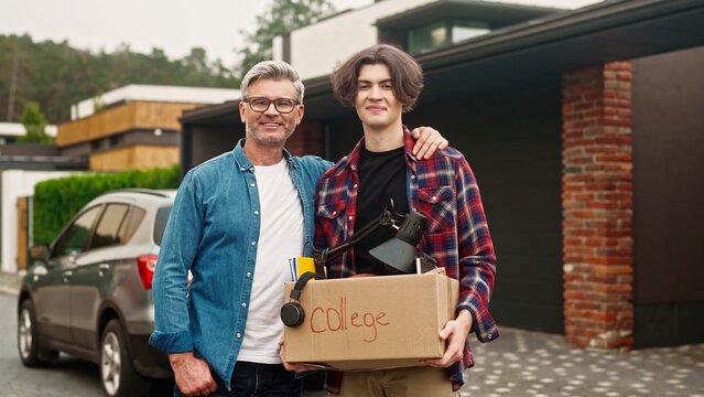 Portrait of father and his son standing outside in yard. Handsome dad hugging his child and proud of his good achievement in college. Satisfied people joyfully smiling while looking on camera.