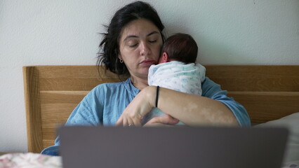 Tired mother holding sleeping newborn baby on her shoulder, sitting in bed with a laptop, depicting the challenges and beauty of balancing work and parenting in daily life