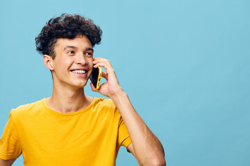 Happy young man talking on smartphone with a bright smile, wearing a yellow t shirt, against a soft blue background, showcasing a cheerful and relaxed lifestyle
