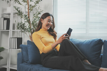 Obraz premium Young businesswoman is sitting on a comfortable couch in her home office, smiling while using her smartphone