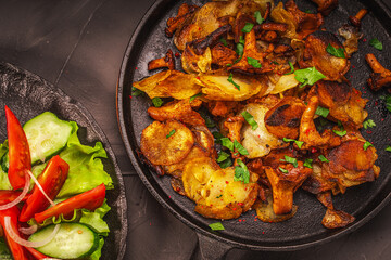 Fried potatoes with chanterelle mushrooms in a cast-iron pan on a dark table with tomatoes and herbs on a plate and lingonberries in a bowl.