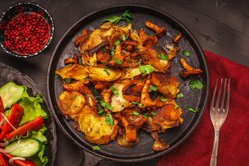 Chanterelle mushrooms fried with potatoes according to a traditional recipe in a cast-iron pan on a dark table with a burgundy napkin, with vegetables and berries