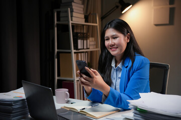 Asian businesswoman is reading a text message on her smartphone and smiling while working late at night in her office