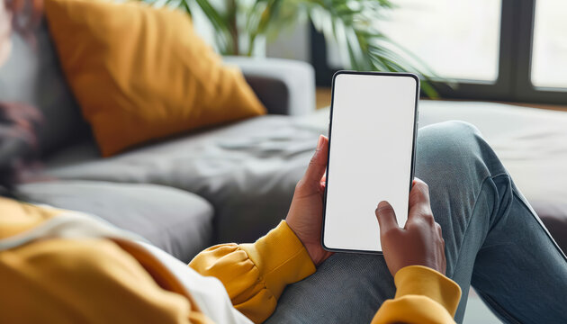 A woman is sitting on a couch holding a cell phone. The phone is white and has a black screen. The woman is focused on her phone, possibly checking messages or browsing the internet