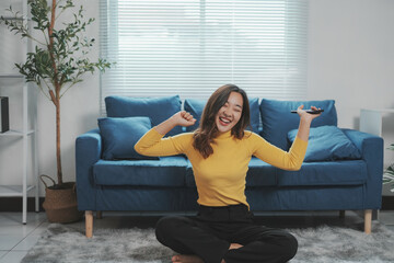 Cheerful young woman is sitting on the floor in the living room, holding a smartphone and dancing while enjoying music