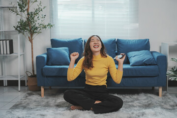 Obraz premium Happy woman sitting on the floor holding a smartphone and raising her arms in the air, celebrating a victory