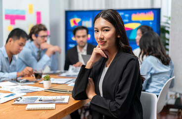 Portrait of happy young asian businesswoman or analyst looking at camera with her colleague analyzing data analysis in dynamic business strategy investment planning meeting. Habiliment