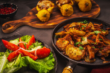 Chanterelle mushrooms fried with potatoes in a cast iron frying pan and stuffed pumpkins on a wooden board, vegetables and a burgundy napkin on a gray table. Traditional autumn dinner.