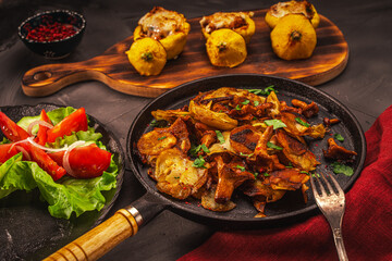 Chanterelle mushrooms fried with potatoes in a cast iron frying pan and stuffed pumpkins on a wooden board, vegetables and a burgundy napkin on a gray table. Festive autumn dinner.