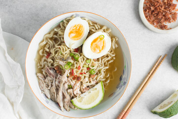 Overhead view of noodles in clear broth with soft boiled eggs, shredded chicken and a lime wedge, top view of ramen noodles in chicken broth on a marble countertop