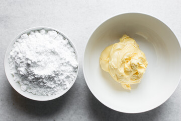 Overhead view of butter and icing sugar in a white mixing bowl, top view of margar and powdered sugar on a marble countertop, ingredients for making buttercream, process of making american buttercream