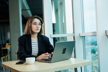 Fototapeta premium A young woman wearing glasses is sitting in a modern spacious working office. Woman office worker with laptop working.