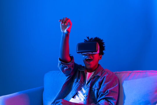 Young African American looking VR enjoy watching fantastic 3D pointing interesting object technology blue neon light virtual reality metaverse new world at living room sitting on sofa. Contrivance.