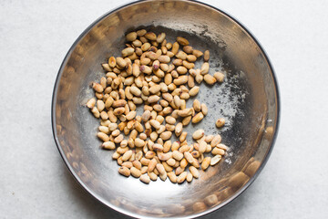 Overhead view of roasted peanuts being coated with egg and flour in a silver bowl, flatlay of roasted peeled peanuts being battered in egg and flour, process of making coated peanuts or cracker nuts