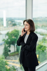 A pretty woman in glasses is standing by the window in a transparent office. A woman rests looking out the window in the office