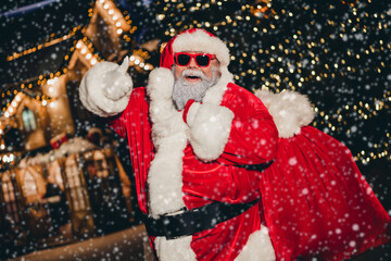 Photo of funky aged man santa costume hold gifts sack demonstrate thumb up feedback fairy atmosphere lights outdoors