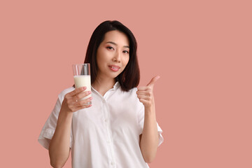 Beautiful Asian woman with glass of milk showing thumb-up on pink background