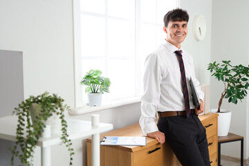 Fototapeta premium Smiling young businessman holding a laptop in a modern office with plants and natural light, the professional male wears a white shirt and tie, exuding confidence and readiness for work