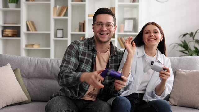 A boy and a girl are having fun playing video games with joysticks in the living room while sitting on the sofa. A couple in love on a date, enjoying the game, joking and having fun.