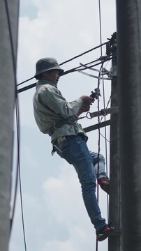 Vietnamese electrician standing on a light pole with a harness repairs electrical cables