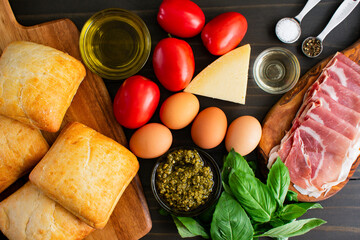 Italian Sandwich Ingredients on Wooden Table: Plum tomatoes, eggs, pesto, ciabatta rolls, parmesan cheese, prosciutto, and basil on dark background