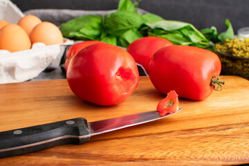 Cored Roma Tomato on a Wooden Cutting Board: Plum tomatoes and paring knife on a wood table with eggs, basil, and a dish of pesto on the background